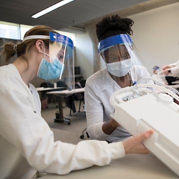 two women wearing PPE masks and white lab coats looking at nursing device